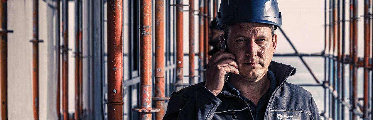 Man met blauwe bouwhelm belt op een bouwplaats terwijl hij tussen steigerelementen staat.