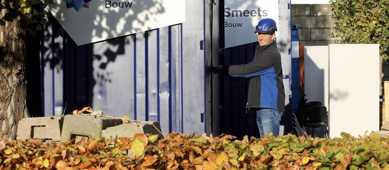 Man met een bouwhelm op pakt iets uit een opslag, met op de voorgrond een heg die verkleurd is door de herfst