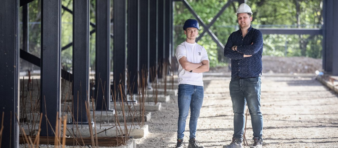 Twee medewerkers met helmen staan met gekruiste armen op een bouwplaats onder een stalen constructie.