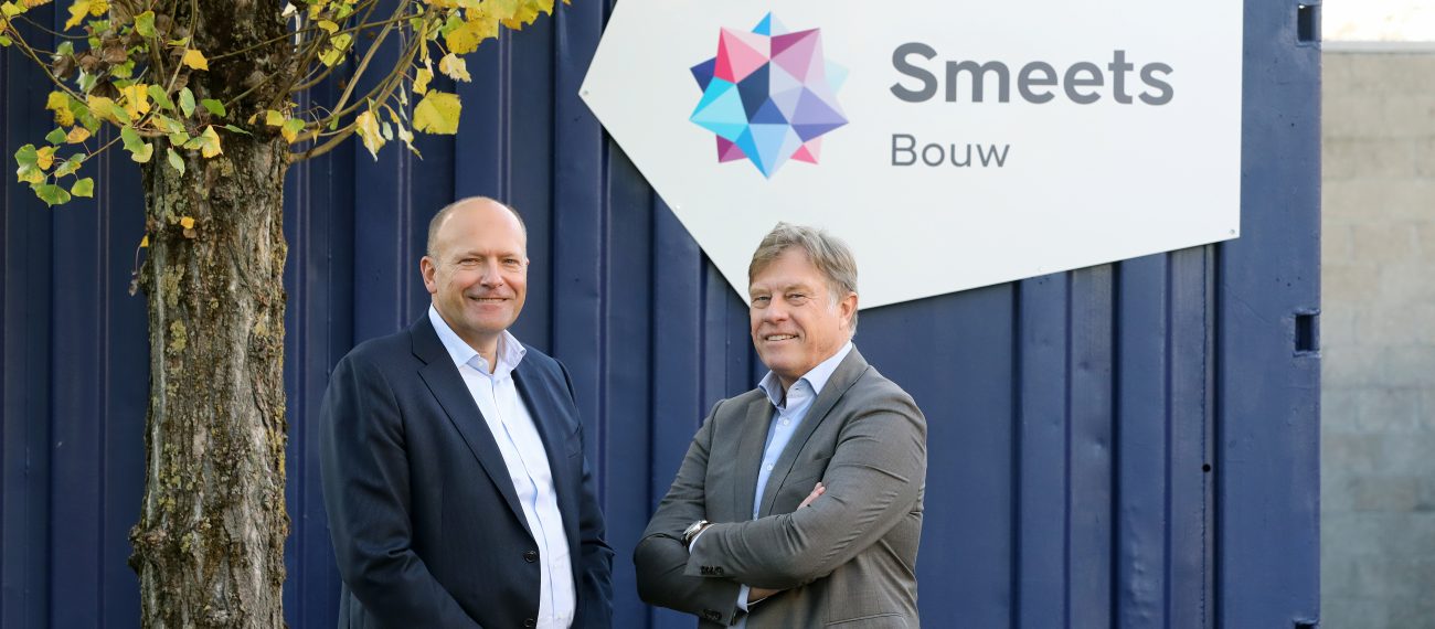 Twee mannen in pak poseren buiten voor een blauw bedrijfsgebouw met boom en een bord met het logo en de naam ‘Smeets Bouw’