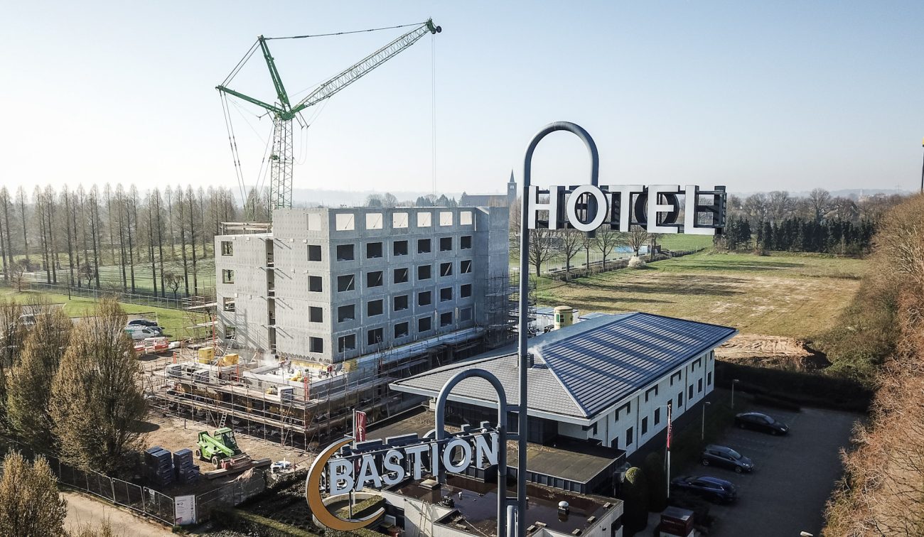 Ruwbouw Bastion Hotel Geleen met betonnen casco en groene torenkraan; op de voorgrond grote “HOTEL”- en “BASTION”-lichtreclames, rechts het lagere bestaande hotel met parkeerplaats, omgeven door bomen en open veld.