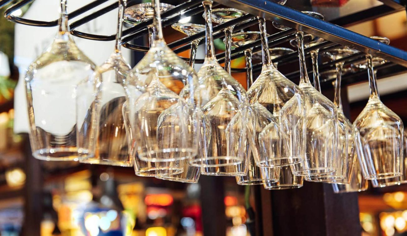Close-up van omgekeerd hangende wijnglazen in een glashouder boven de bar, met warme caféverlichting op de achtergrond (Eetcafé De Aanwinst, Nederweert).