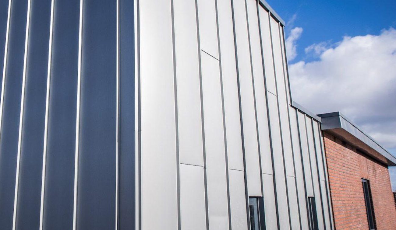 Detail van gevel met verticale metalen felsbanen in grijstinten naast een rood­bakstenen wand; smalle raampartijen, onder blauwe lucht met wolken.