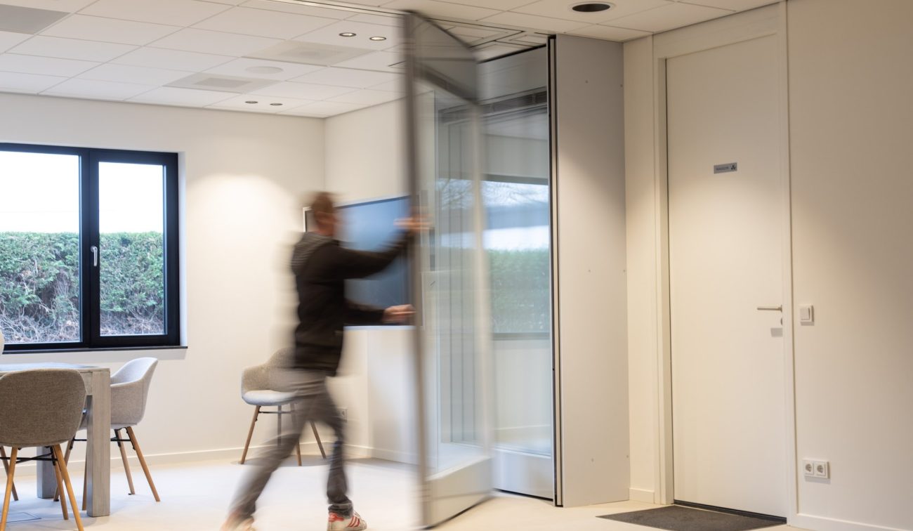 Interieur kantoor met een beweegbare glazen/akoestische vouwwand die door een persoon in beweging wordt dichtgeschoven; links vergaderhoek met stoelen en raam, rechts een witte deur.