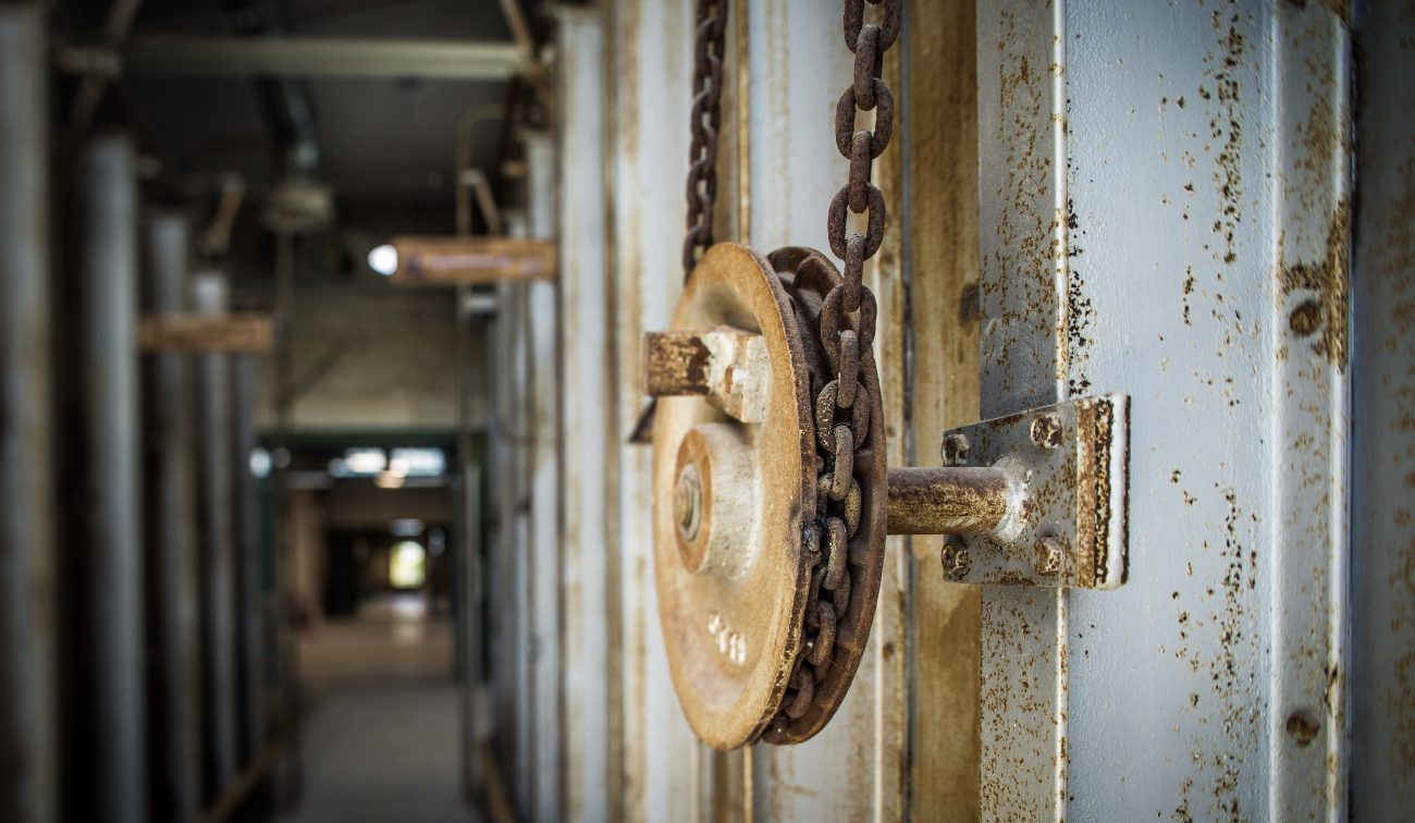 Close-up van een roestige katrol met ketting aan een stalen balk; industriële hal op de achtergrond onscherp.