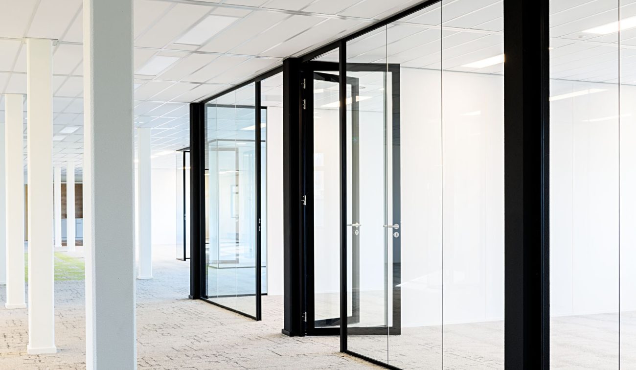 Modern kantoor bij zorginstelling: glazen kamers, stalen kozijnen en lichte tapijttegels op vloer