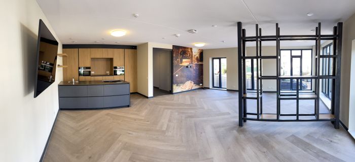Interieur zorgcomplex Cicero Bleekstaete: ruime woonkamer met visgraatvloer, ingebouwde houtlook keuken, zwarte open vakkenkast als roomdivider, grote ramen en dubbele deuren naar balkon; wand met fotoprint en plafondspots.