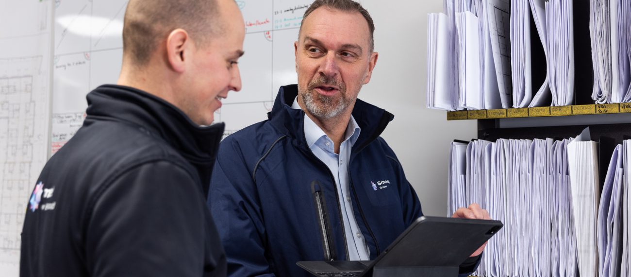 Twee collega’s overleggen in een kantoorruimte; één houdt een tablet vast terwijl ze voor een wand met ordners staan.
