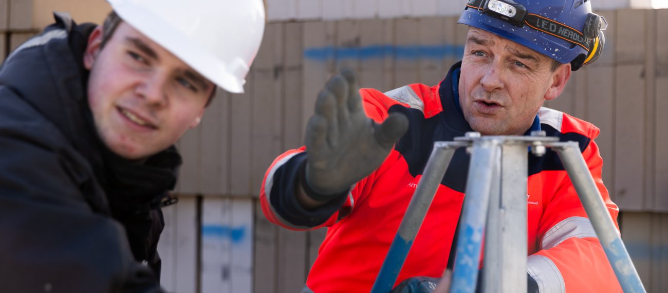 Twee mannen met helmen werken samen op een bouwplaats; één geeft aanwijzingen terwijl de ander een statief vasthoudt.