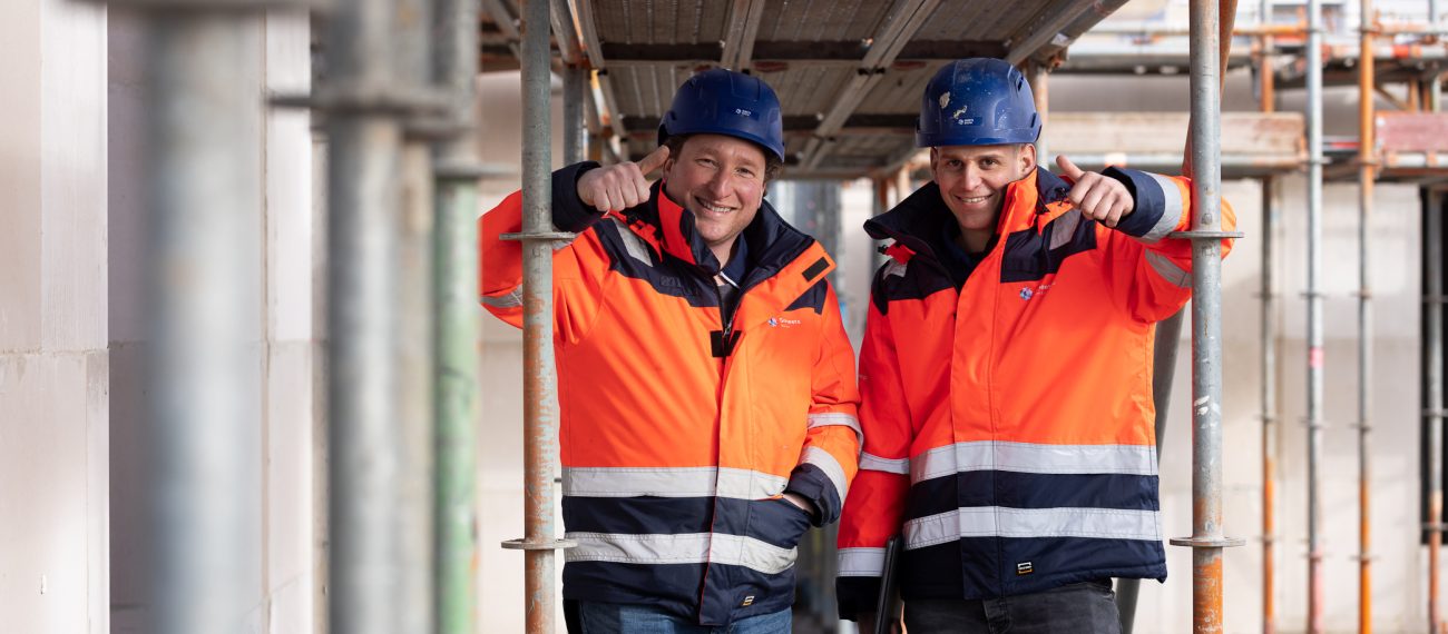 Twee medewerkers in bouwkleding geven een duim omhoog op een bouwplaats