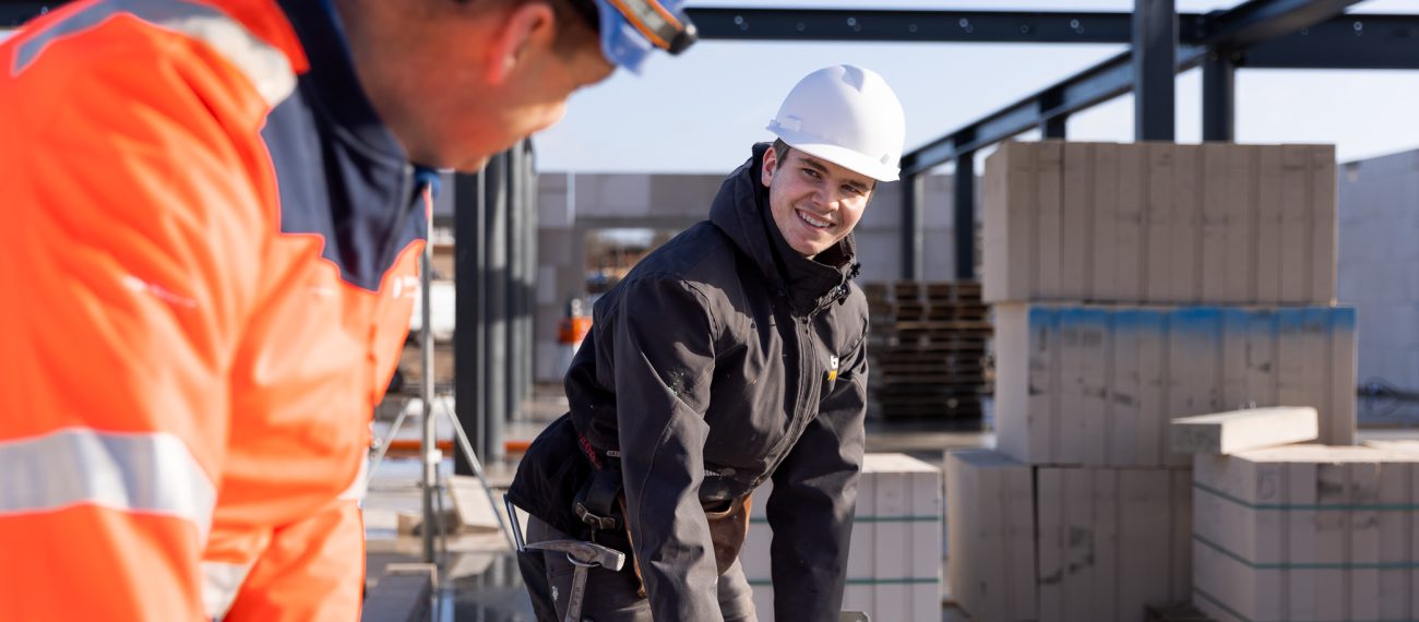 Twee medewerkers op een bouwplaats met helmen en werkkleding; één persoon tilt een buis en glimlacht naar een collega, met bouwmaterialen en staalconstructie op de achtergrond.”