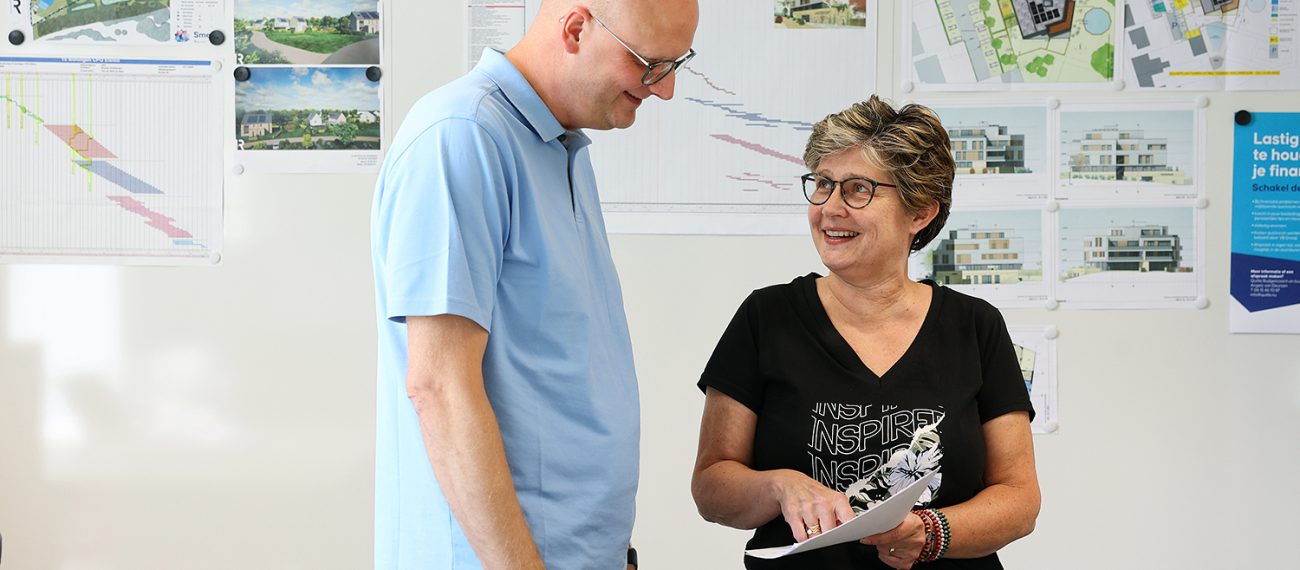 Twee collega’s bespreken bouwtekeningen in kantoor, met projectvisuals en plattegronden op de muur.