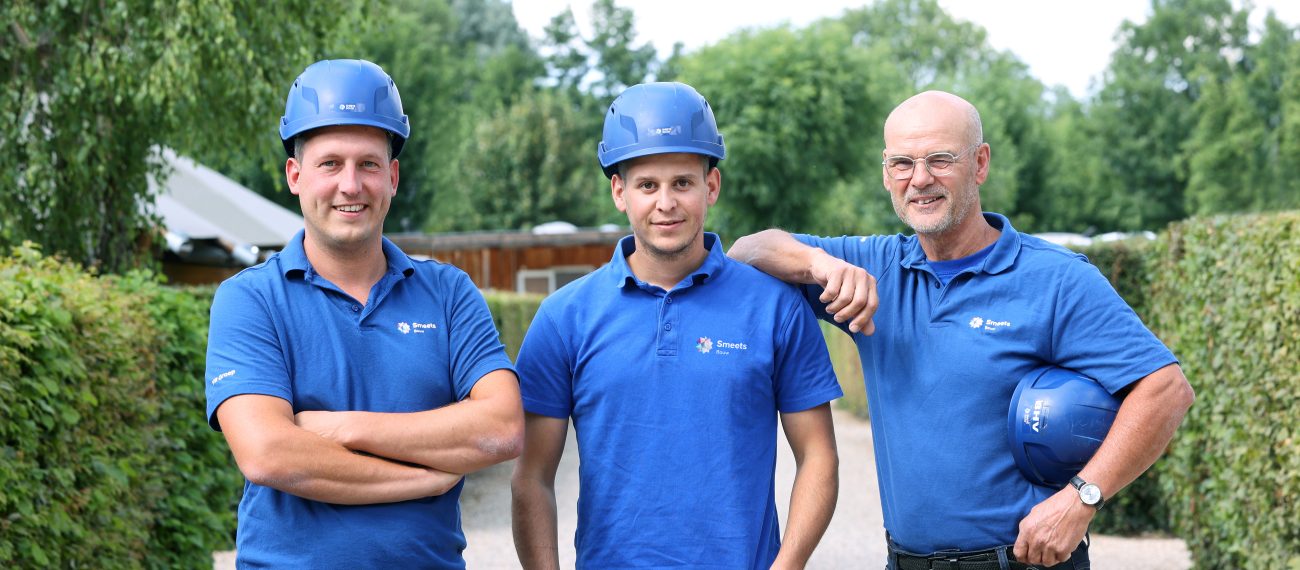 Drie medewerkers in blauwe bedrijfskleding en helmen poseren buiten op een pad, glimlachend naar de camera.