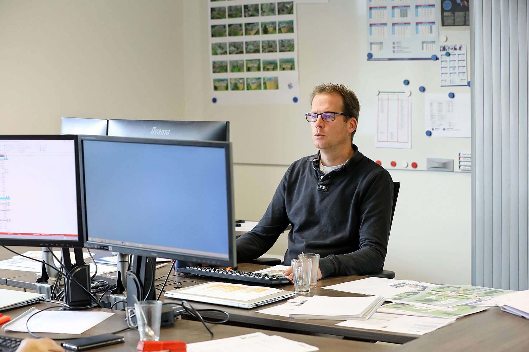 Medewerker achter twee beeldschermen aan een kantoorwerkplek, omringd door bouwtekeningen en documenten