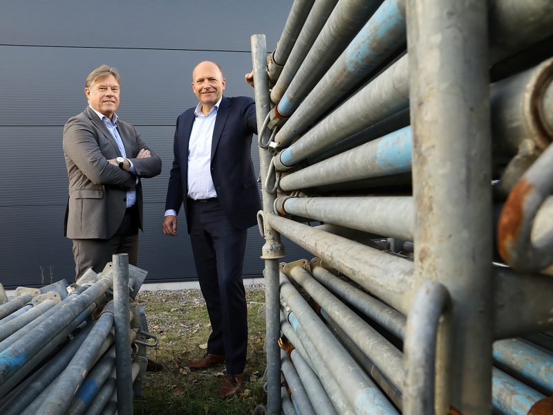 Twee mannen in pak staan buiten bij een wand van metalen steigerbuizen en kijken lachend naar de camera