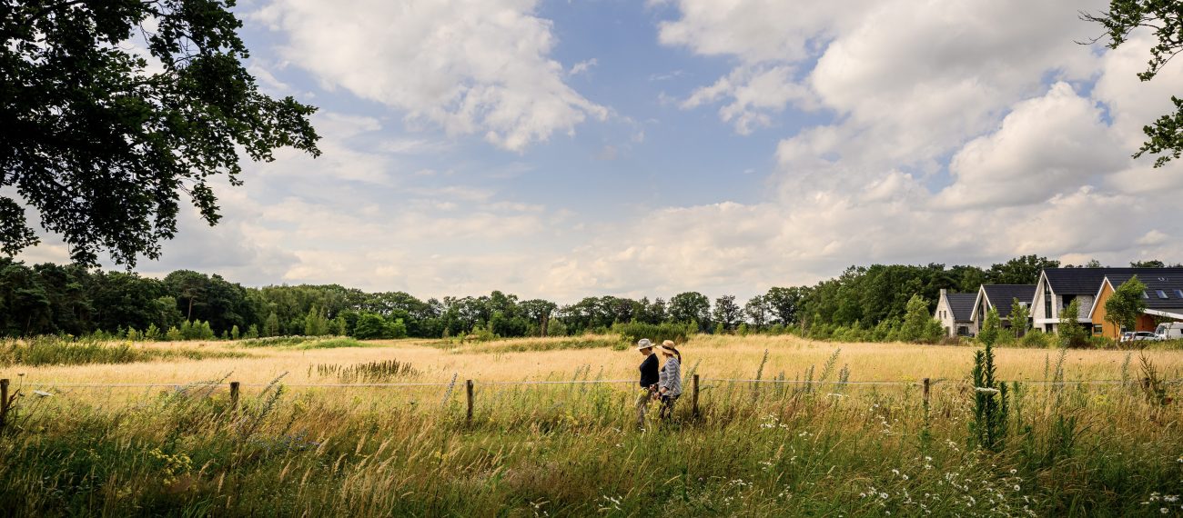 Twee vrouwen wandelen in een veld in een groene omgeving met blauwe lucht en bewolking, met een aantal huizen op de achtergrond