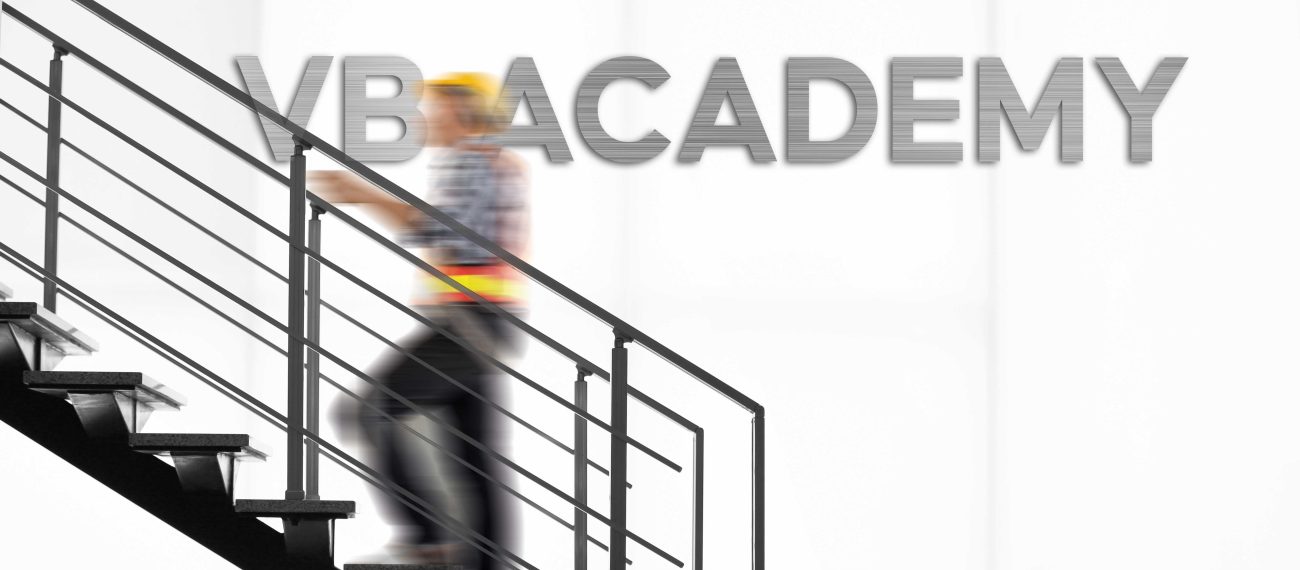 Vage figuur van medewerker met veiligheidshelm loopt de trap op voor een witte wand met de tekst ‘VB ACADEMY’ erboven.