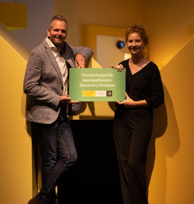 Twee mensen poseren met een bord waarop staat "Maatschappelijk Aandeelhouder Discovery Mseum"