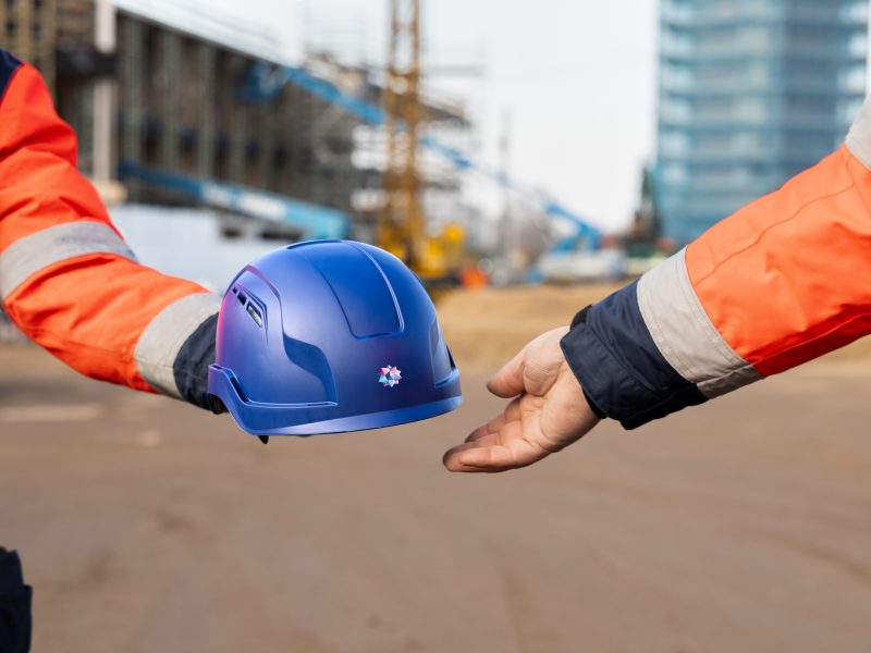 Iemand geeft een bouwhelm aan een collega, waarbij op de achtergrond twee gebouwen in de steigers staan