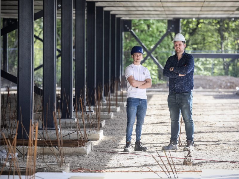 Twee medewerkers met helmen staan met gekruiste armen op een bouwplaats onder een stalen constructie.