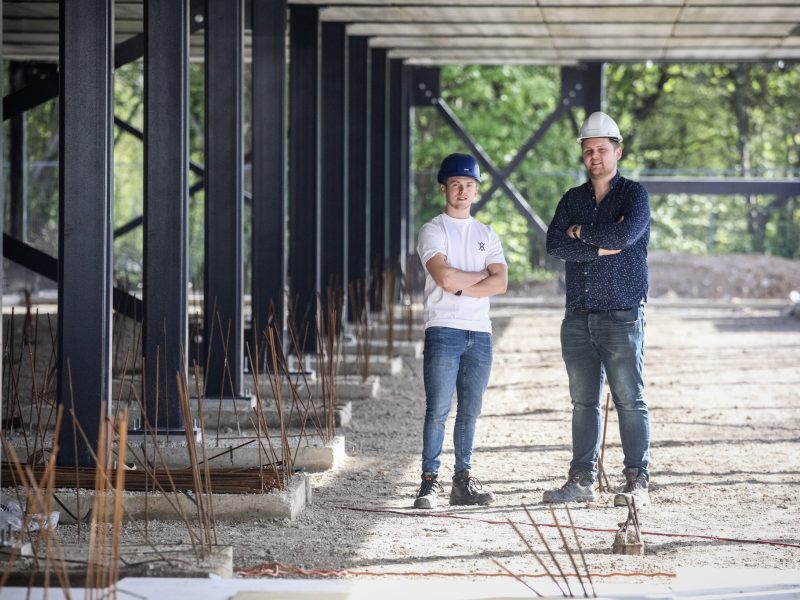 Twee personen met veiligheidshelmen staan met gekruiste armen op een bouwplaats tussen stalen kolommen en werkmateriaal.