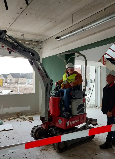 Minigraver tijdens sloopwerk in binnenruimte met bestuurder in veiligheidshelm en hesje