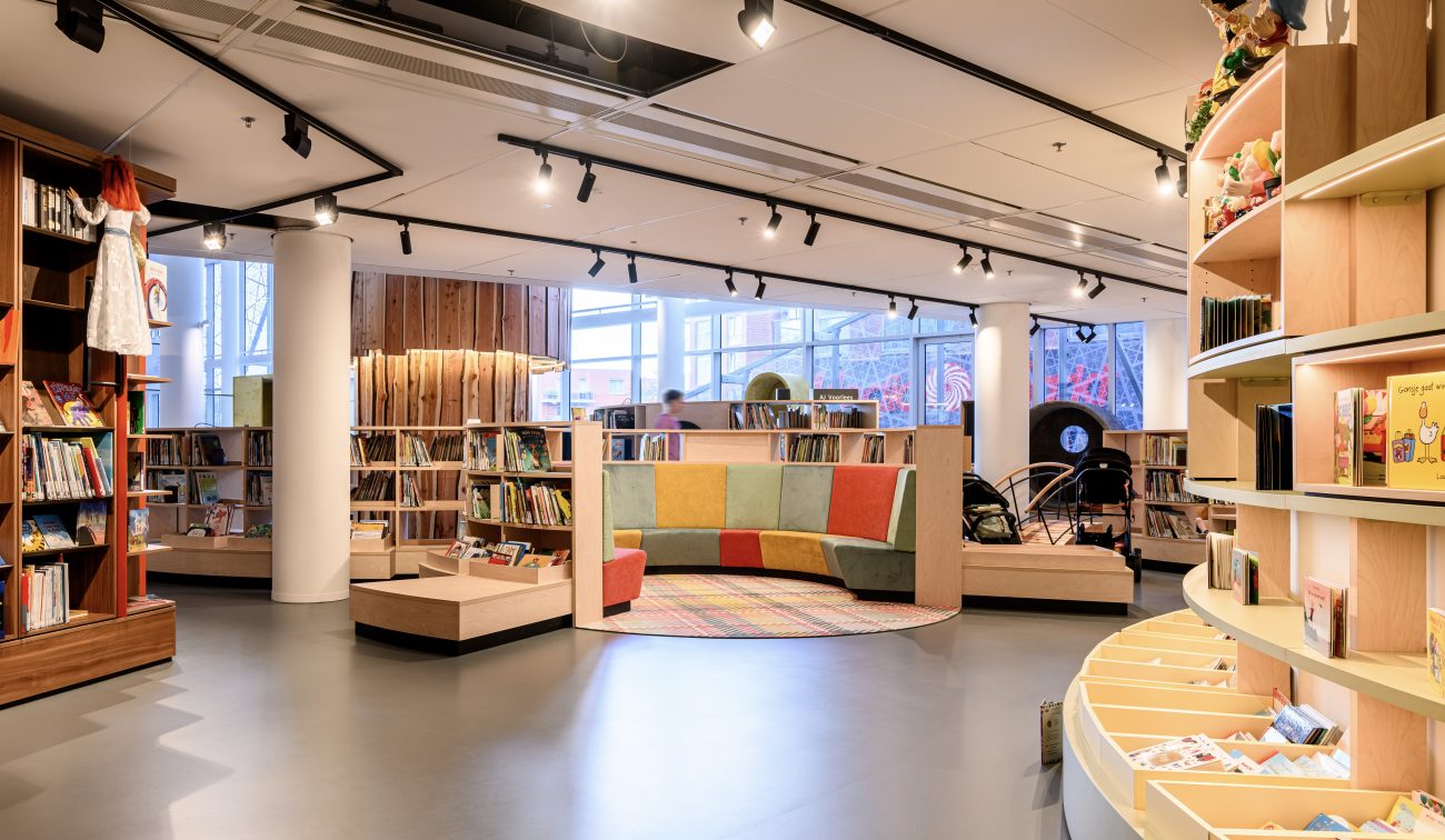 Kinderruimte in bibliotheek met halfronde gekleurde bank, lage boekenkasten en spotverlichting.
