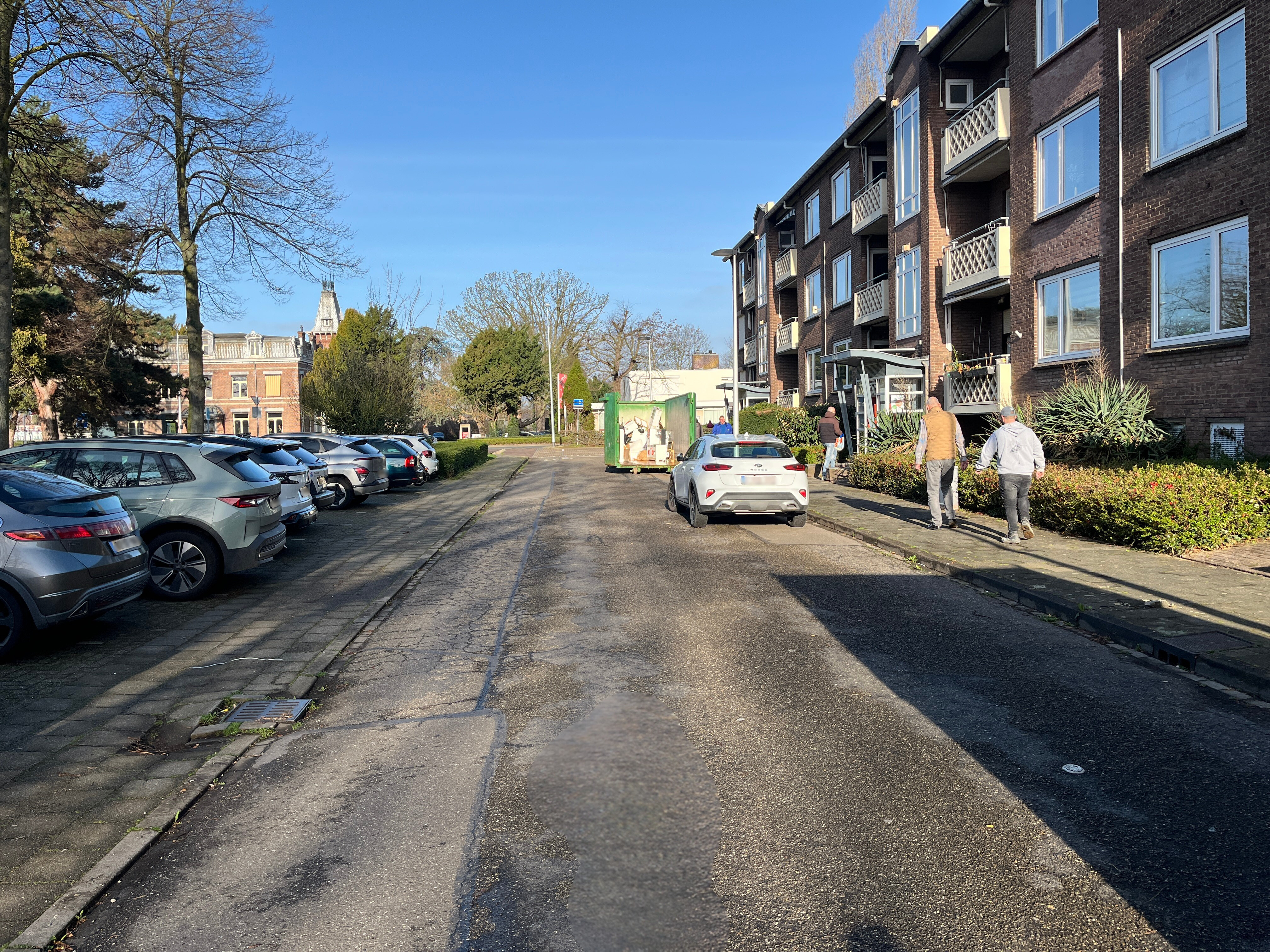 Een straat met aan de rechterzijde appartementen en aan de linkerzijde een parkeerplaats met auto's