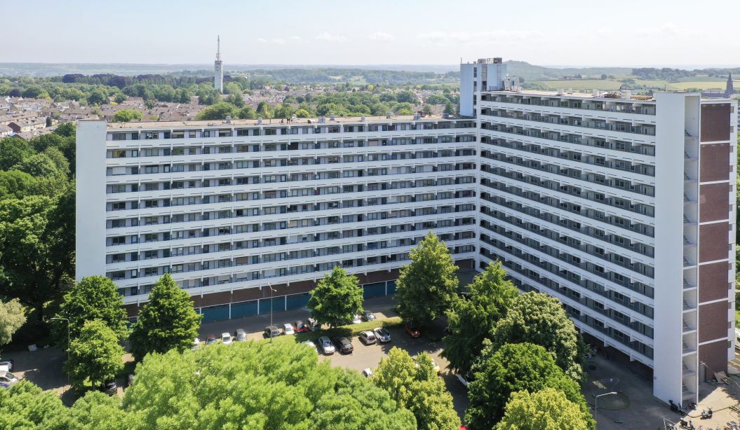 Luchtfoto van L-vormige galerijflat Veliahof in Maastricht, met witte gerenoveerde gevels en lange balkonstroken, omgeven door bomen en parkeerplaatsen.