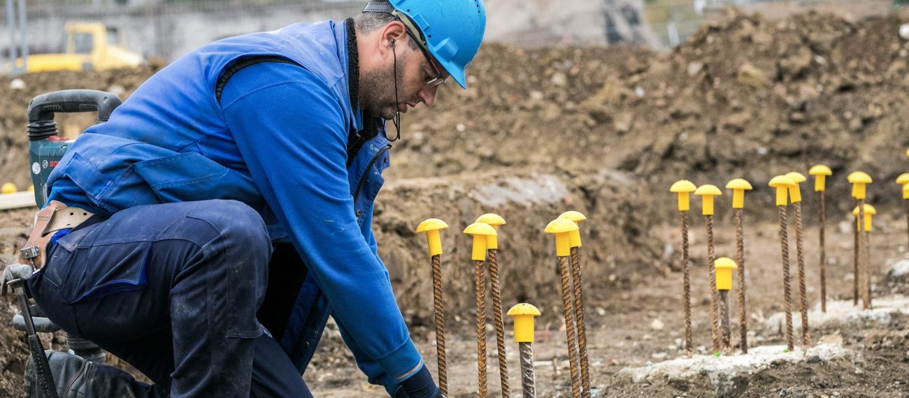 Bouwvakker met blauwe helm en werkkleding werkt op een bouwplaats tussen uitstekende wapeningstaven met gele veiligheidsdoppen in een ruige, onverharde omgeving.