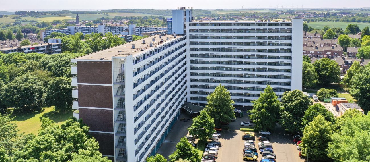 Luchtfoto van L-vormige galerijflat Veliahof in Maastricht met witte gerenoveerde gevels en lange balkonstroken, omringd door bomen en parkeerplaatsen.