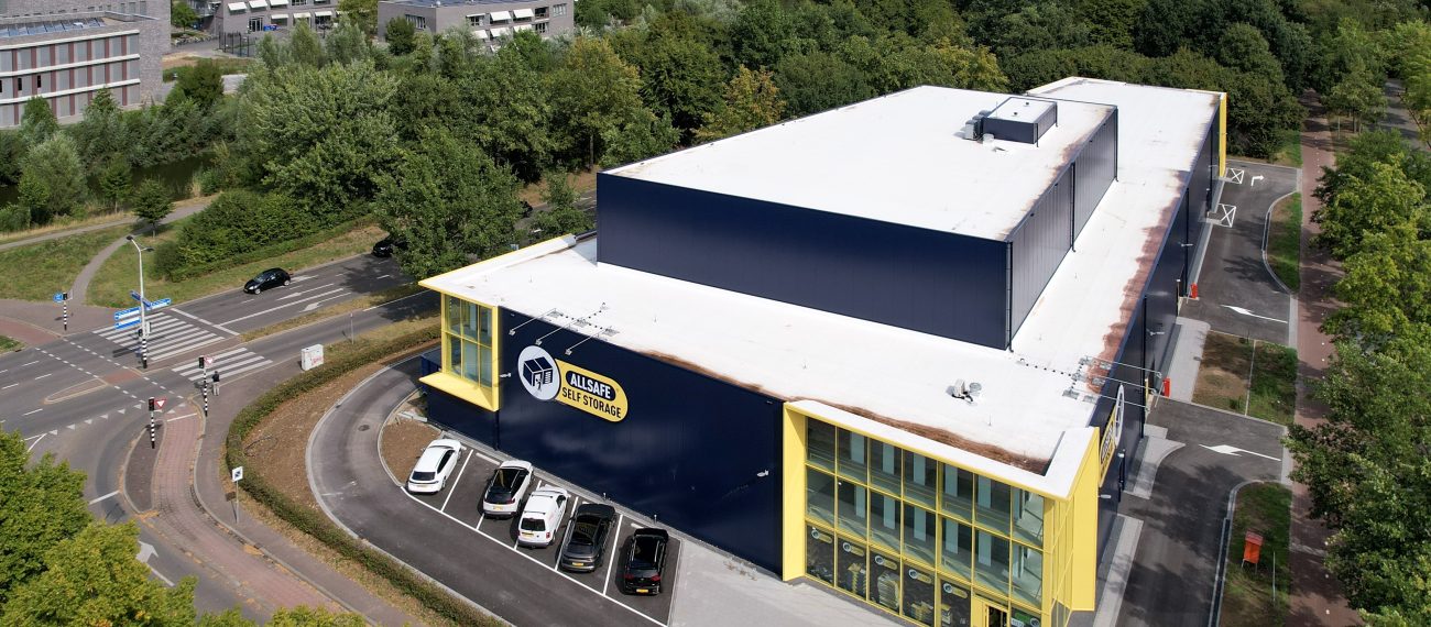 Luchtfoto van de nieuwbouw van Allsafe Self Storage in Maastricht: donkerblauw gebouw met witte dakplaat en felgele kaders rond de glazen hoekgevel, kleine parkeerstrook met auto’s langs de gevel, gelegen aan een kruising en omringd door bomen.