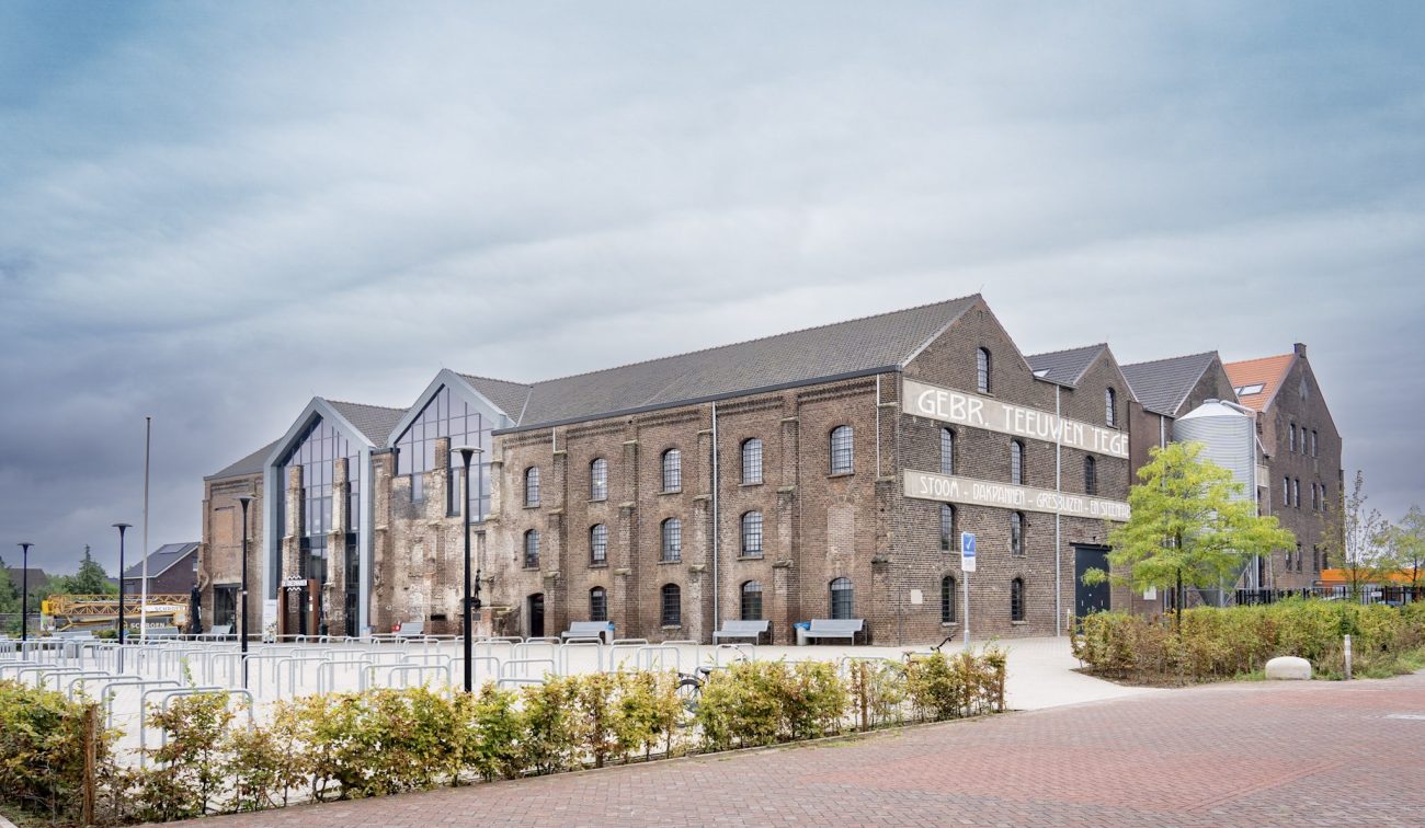 Gerestaureerde Greswarenfabriek in Reuver met bakstenen gevels, grote glazen entree en historische gevelletters; voorliggend plein met fietsrekken, bankjes en jonge bomen.