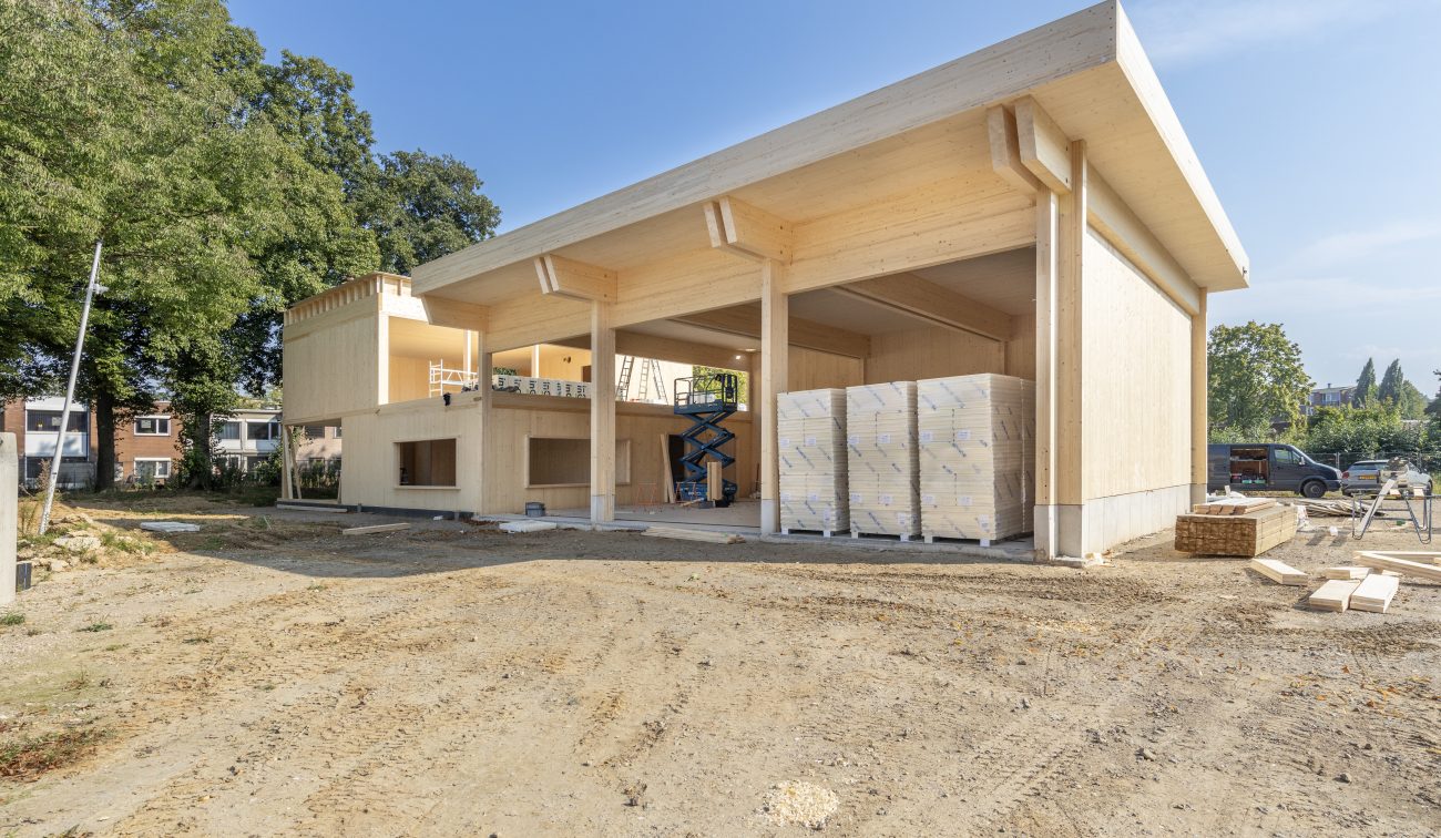 Bouwfase van de nieuwbouw brandweerpost Sittard-Oost: houten casco met grote overkapping en open voertuighal, glulam kolommen/balken, stapels bouwmateriaal en een schaarlift op een zandig bouwterrein onder blauwe lucht.