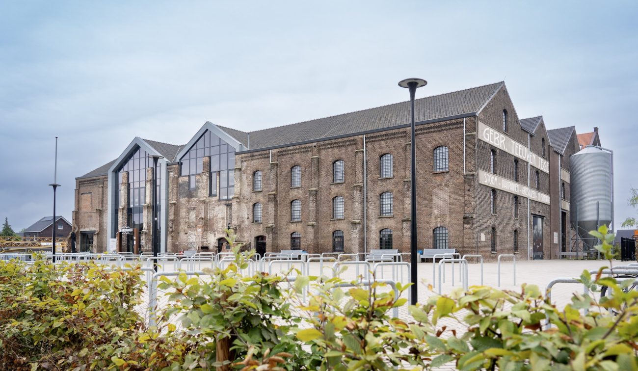 Getransformeerde Greswarenfabriek in Reuver: gerestaureerd bakstenen fabriekspand met grote glazen entree, oude gevelletters en silo aan het plein met fietsenrekken en lantaarnpalen.