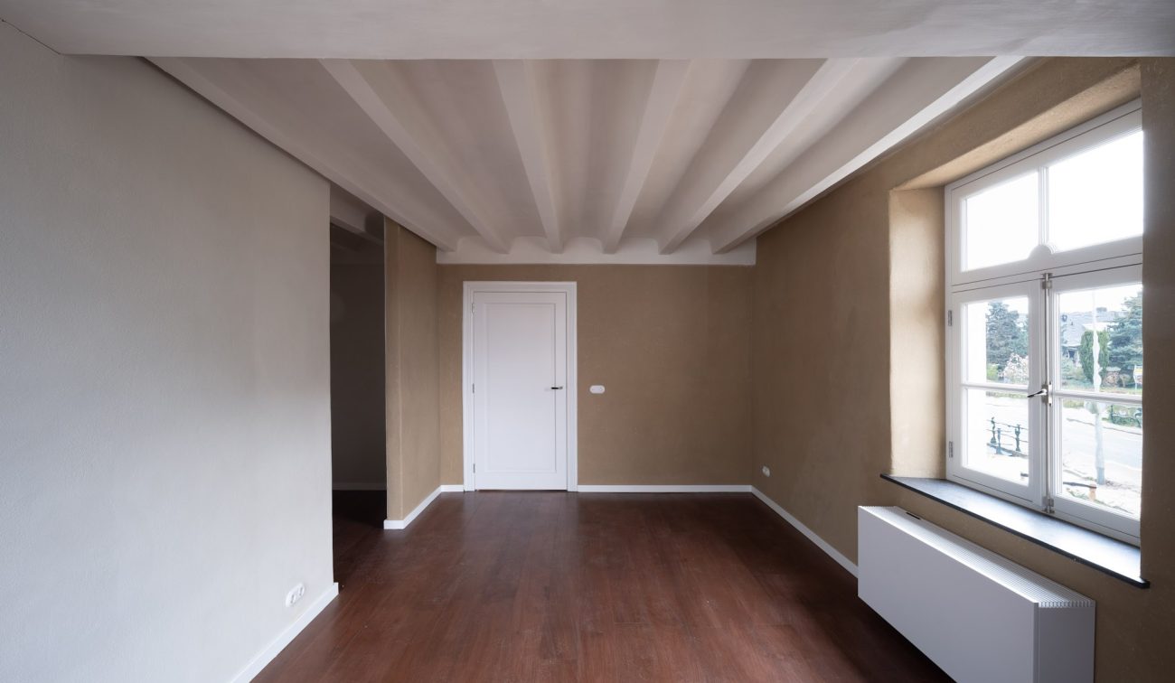 Gerestaureerde kamer met houten vloer, wit balkenplafond, beige stucwanden, een witte binnendeur en een groot raam met vensterbank en radiator, uitkijkend op de omgeving van de Grathemermolen.