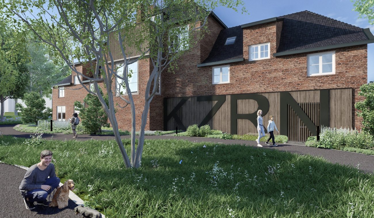 Impressie van de nieuwbouwkazerne in Helmond overdag, met een modern gebouw van rood metselwerk en houten gevel met grote letters ‘KZRN’, omgeven door groen en wandelpaden waar mensen en huisdieren lopen.