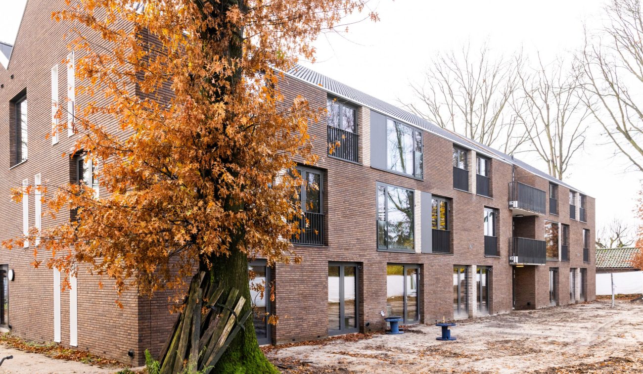 Nieuwbouw van woonzorgcomplex Pontenage in Gemert, een modern bakstenen gebouw met grote ramen en balkons, omgeven door bomen met herfstbladeren
