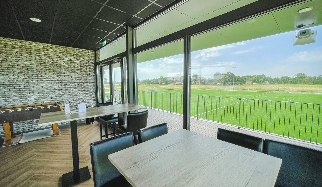 Kantine DVO’20 met hoge en lage tafels bij grote raampartijen, uitzicht op het voetbalveld en terras met balustrade; bakstenen wand en visgraatvloer.