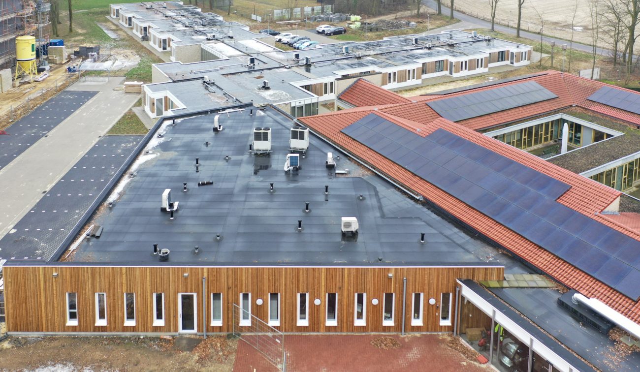 Luchtfoto nieuwbouw zorgwoningen Amstenrade: paviljoen met houten gevel, zwart plat dak met installaties, naast rood pannendak met grote zonnepanelen.