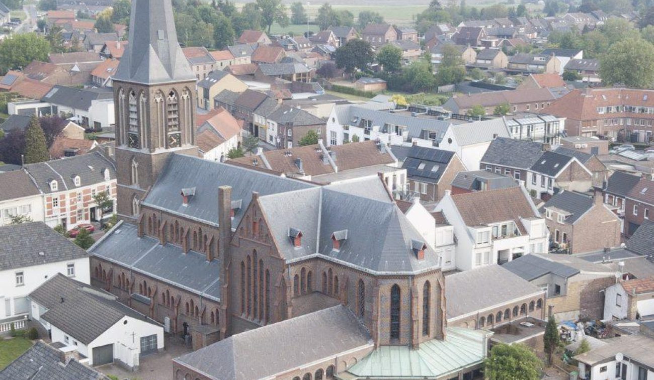 Luchtfoto bakstenen kerk met hoge torenspits, kloostergang en zinken daken midden in dorpsker