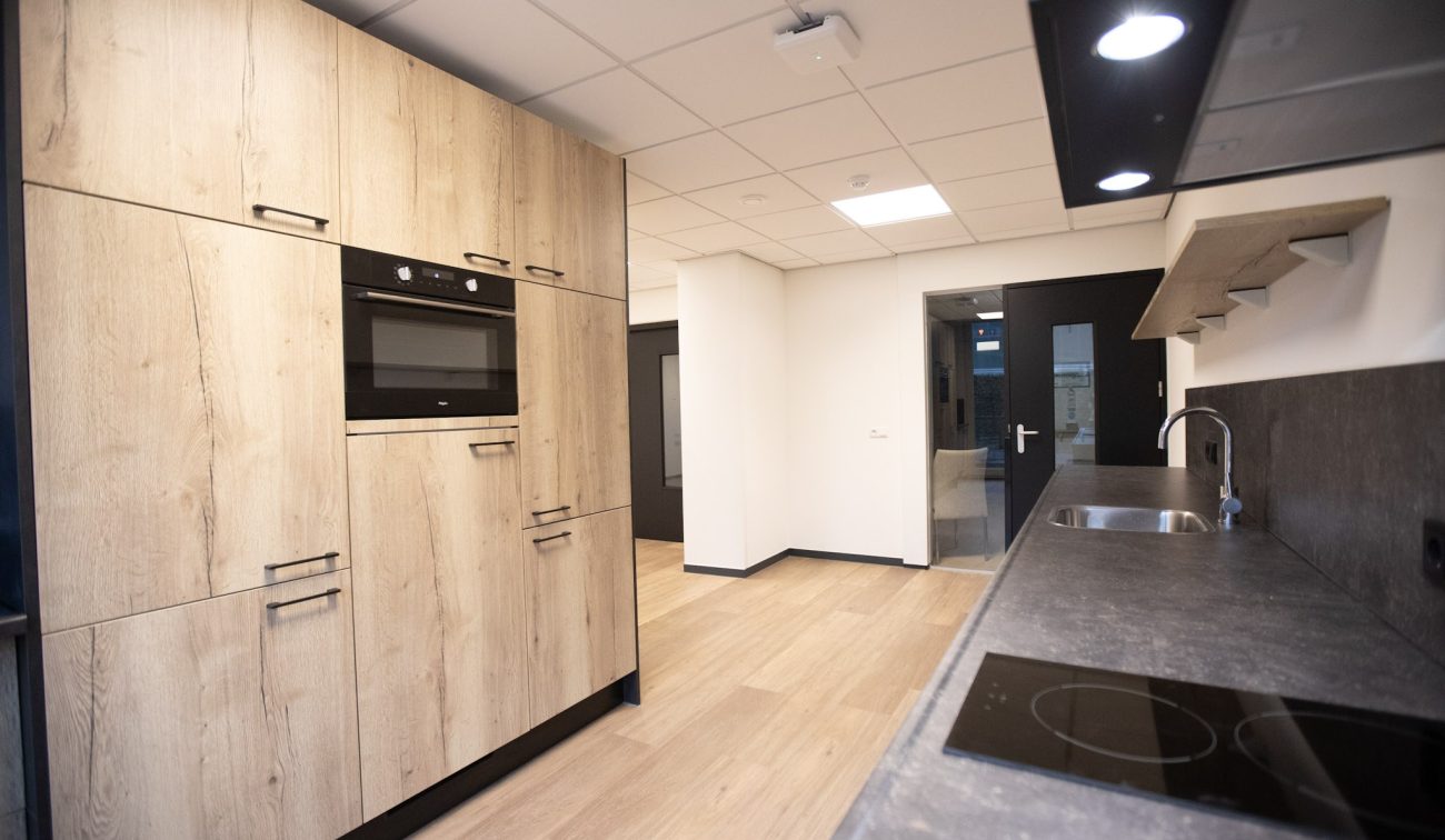 Moderne kitchenette met houtlook kastenwand, inbouwoven, donker werkblad met spoelbak en inductiekookplaat; witte wanden en plafonddpanelen, zicht op aangrenzende gang en deuren.