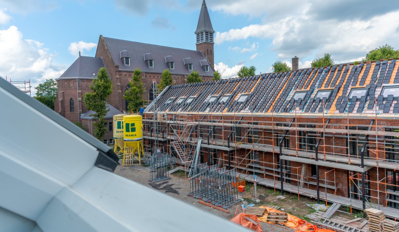 Kloostervelden Sterksel – rijwoningen in aanbouw met steigers en dakpannenmontage, gele silosilo; kerktoren op achtergrond