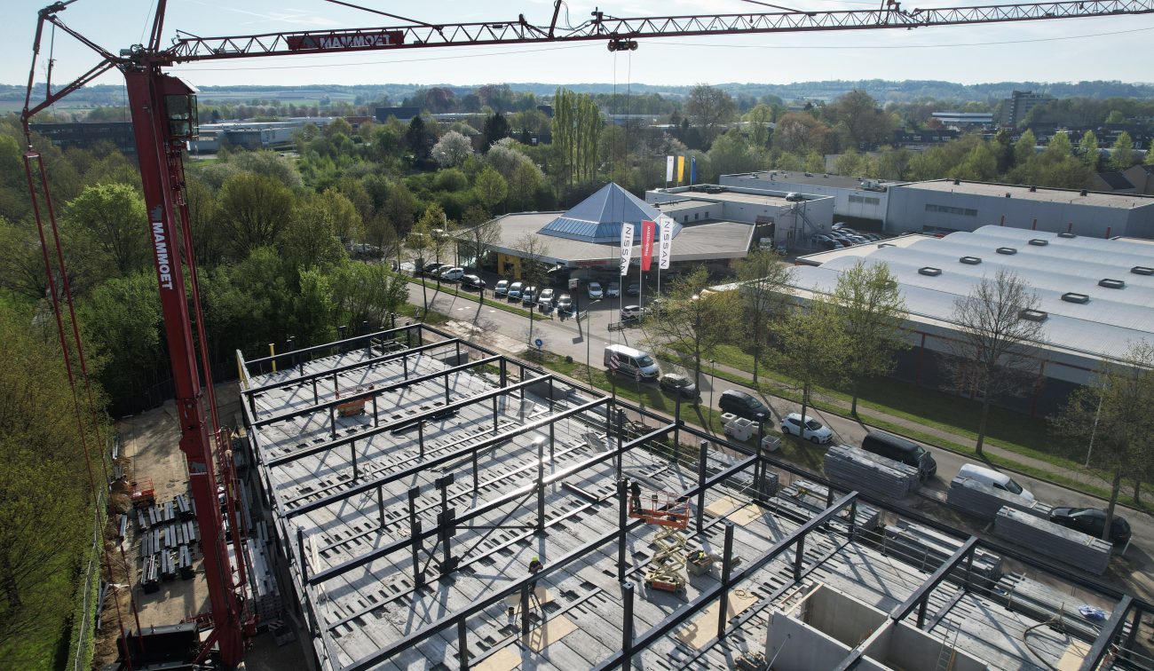 Schuine dronefoto van een bouwplaats met stalen skelet en kanaalplaatvloeren; links een rode kraan, op de vloer werken twee bouwvakkers. Aan de overkant een straat met auto’s en bedrijfsgebouwen, waaronder een showroom met blauw piramidedak.