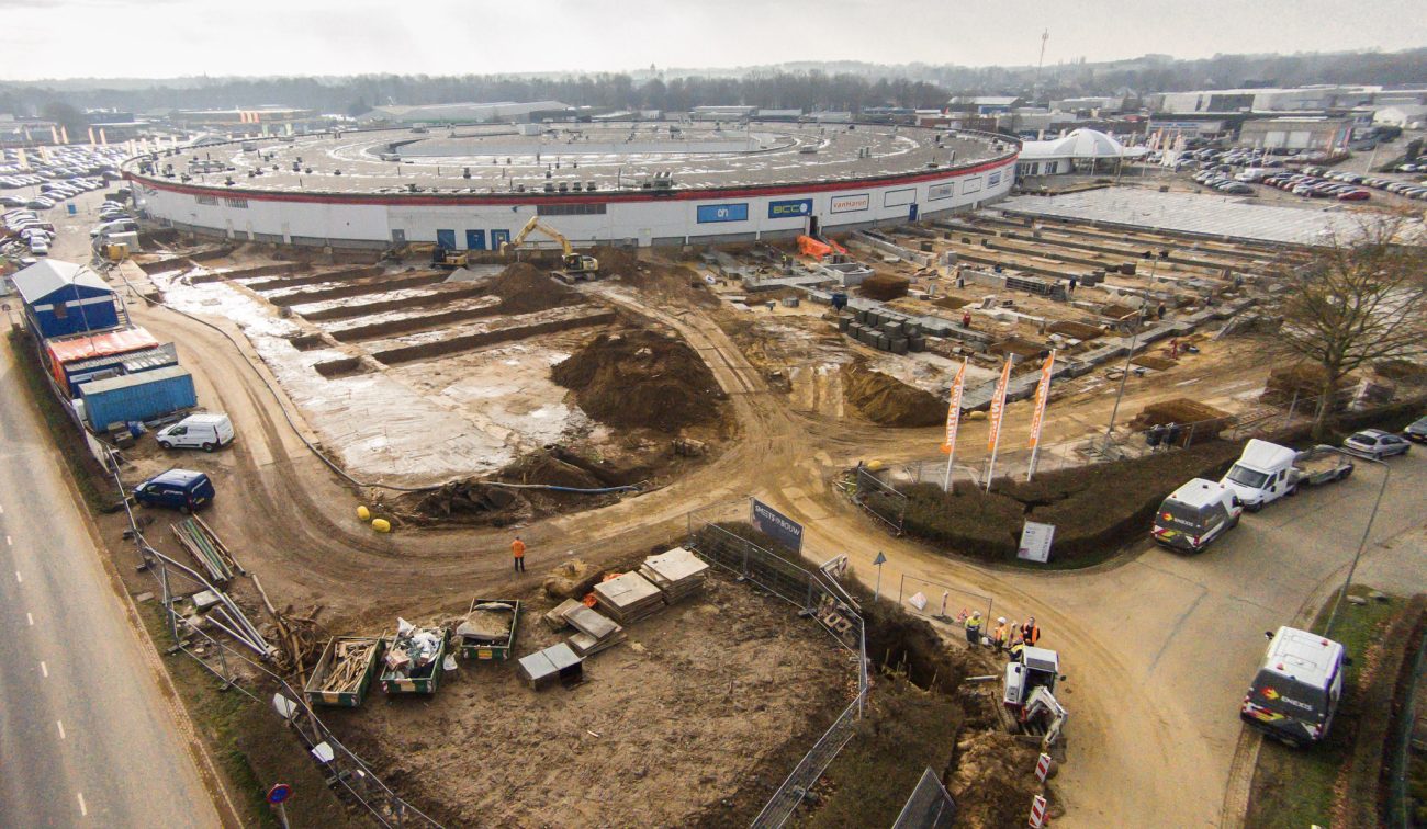 Luchtfoto uitbreiding Makado Beek met bouwplaats, funderingen en winkelcentrum op achtergrond.