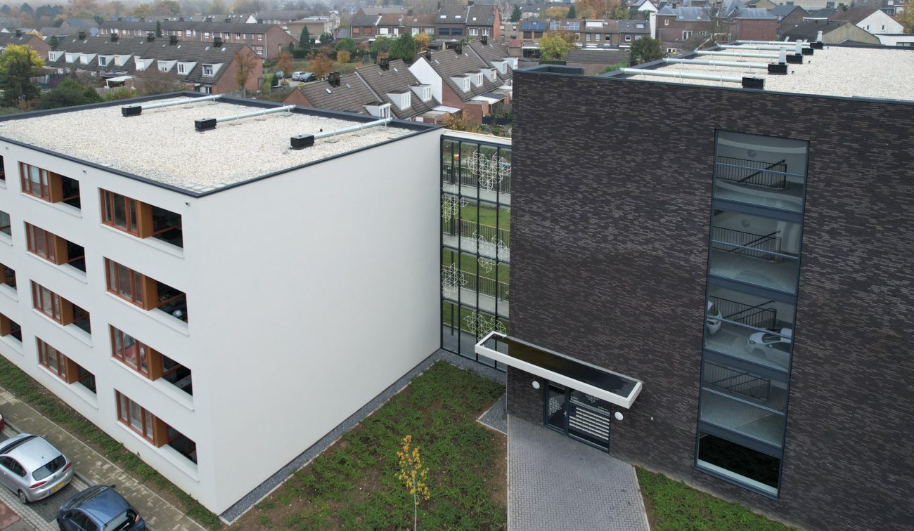 Nieuwbouw appartementen Magistraat Heerlen: wit gestuct woonblok gekoppeld aan een donker bakstenen gebouw met glazen trappenhuis en een entree met zwarte luifel.