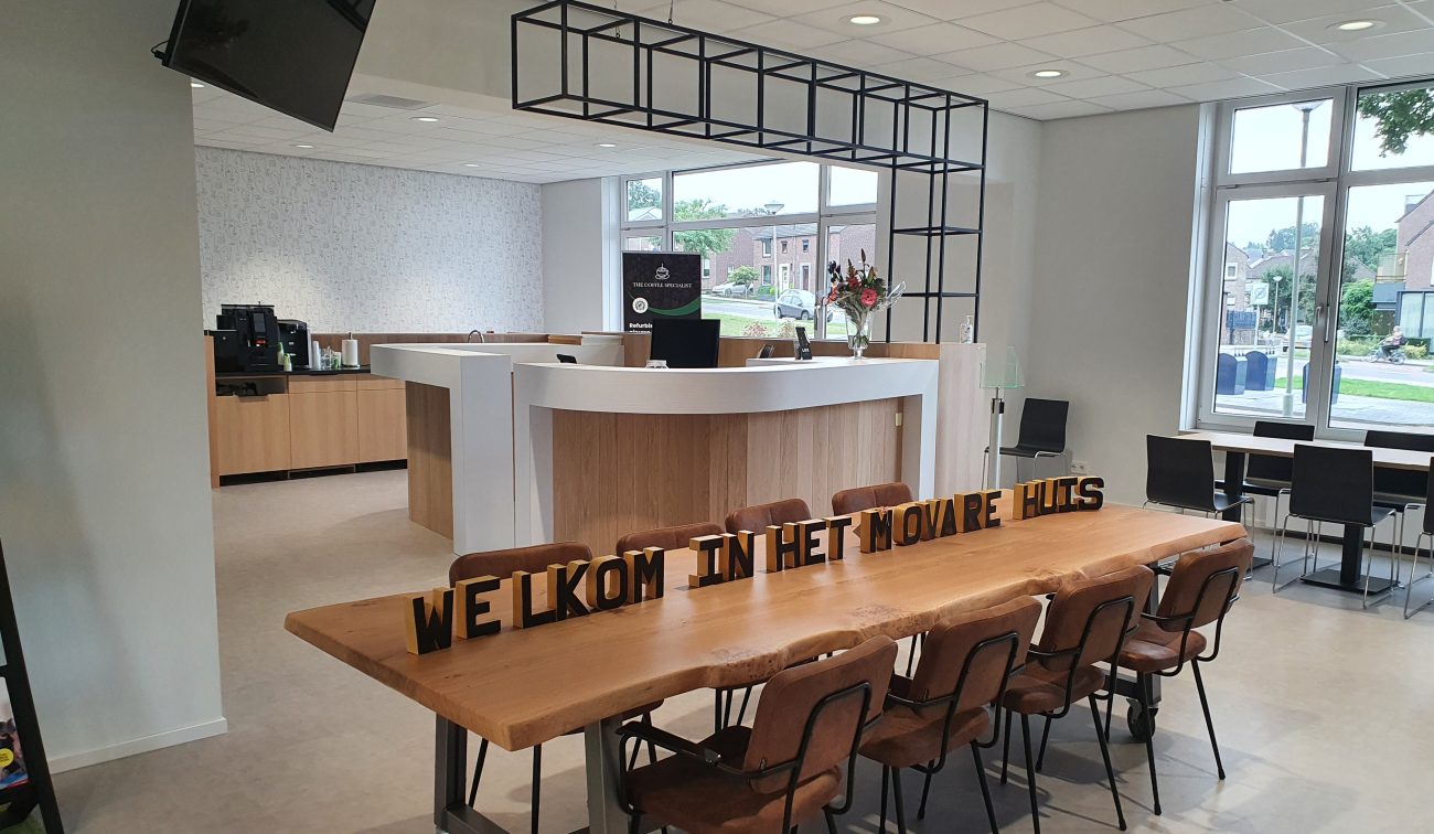 Moderne ontvangstruimte met witte balie en koffiecorner; centraal staat een lange houten tafel met stoelen en de tekst ‘WELKOM IN HET MOVARE HUIS’. Grote ramen en plafondspots zorgen voor veel licht.