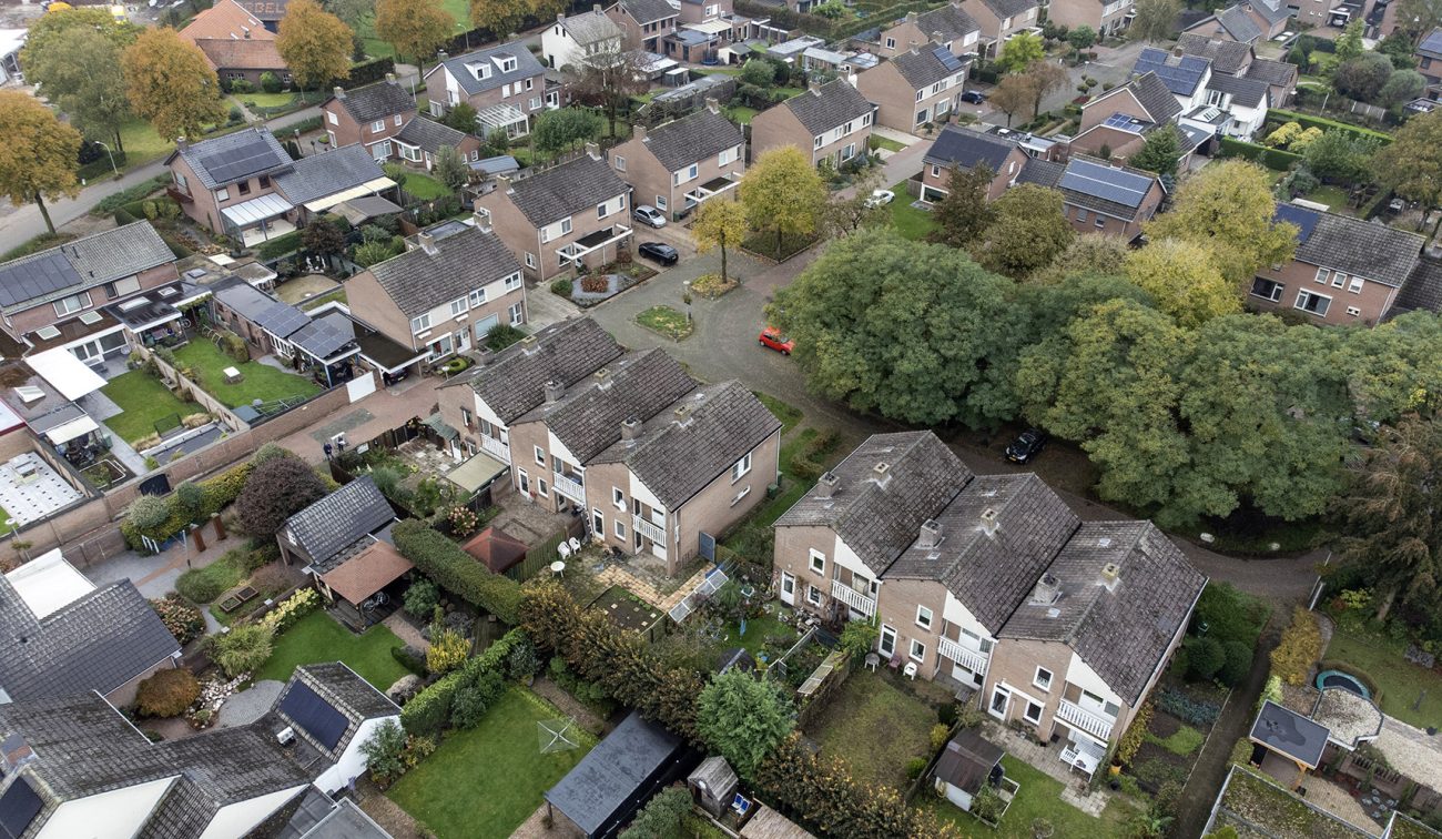 Luchtfoto van een woonbuurt in Stramproy: rijen rijwoningen en twee-onder-een-kap met achtertuinen, veel groen en enkele zonnepanelen—locatie voor groot onderhoud aan 28 woningen.