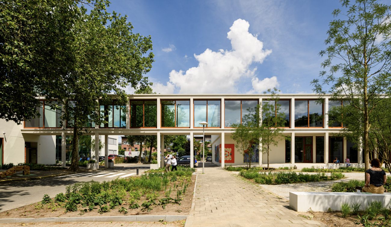 Transparant schoolgebouw met loopbrug en glas; studenten wandelen tussen het groen.