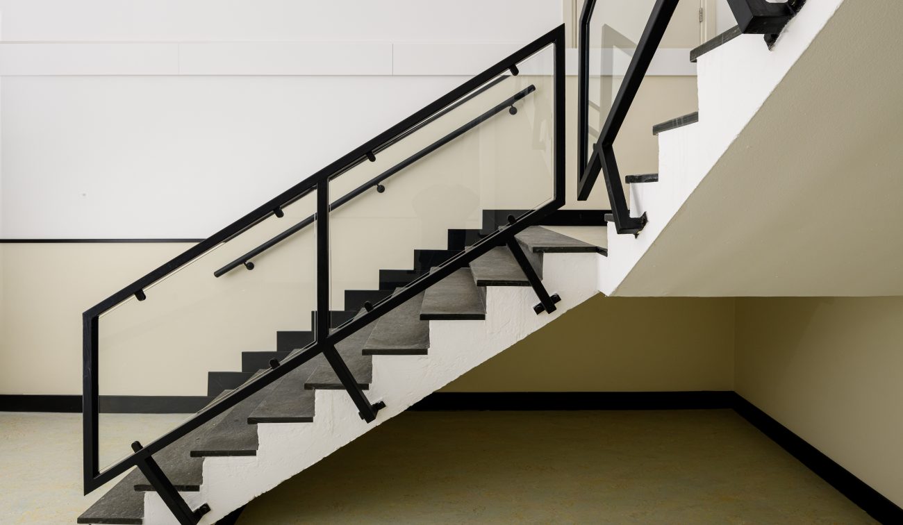 Moderne schooltrap: glaspanelen, dubbele handregel langs de muur en grijze steen-/rubbertreden.